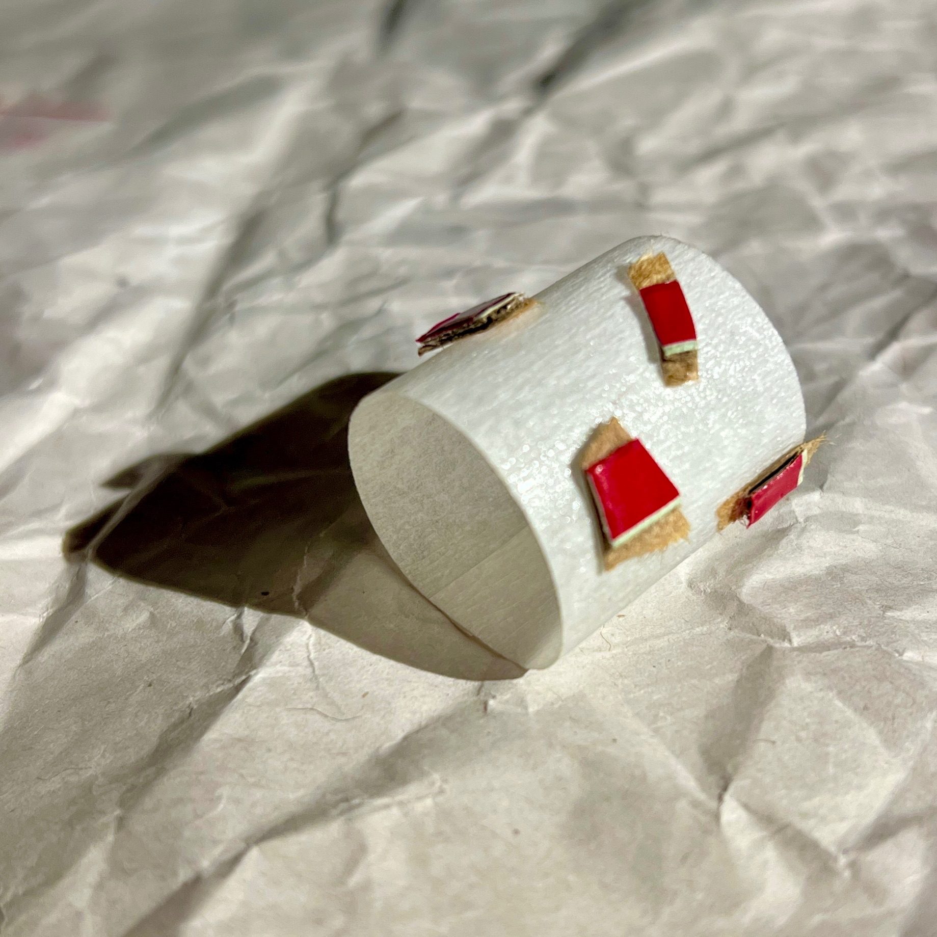 A small tube of masking tape, dotted with tiny pieces of rip-strip from a cardboard envelope sits on a piece of crumpled paper. A strong light from the right casts a shadow to the left of the tube.