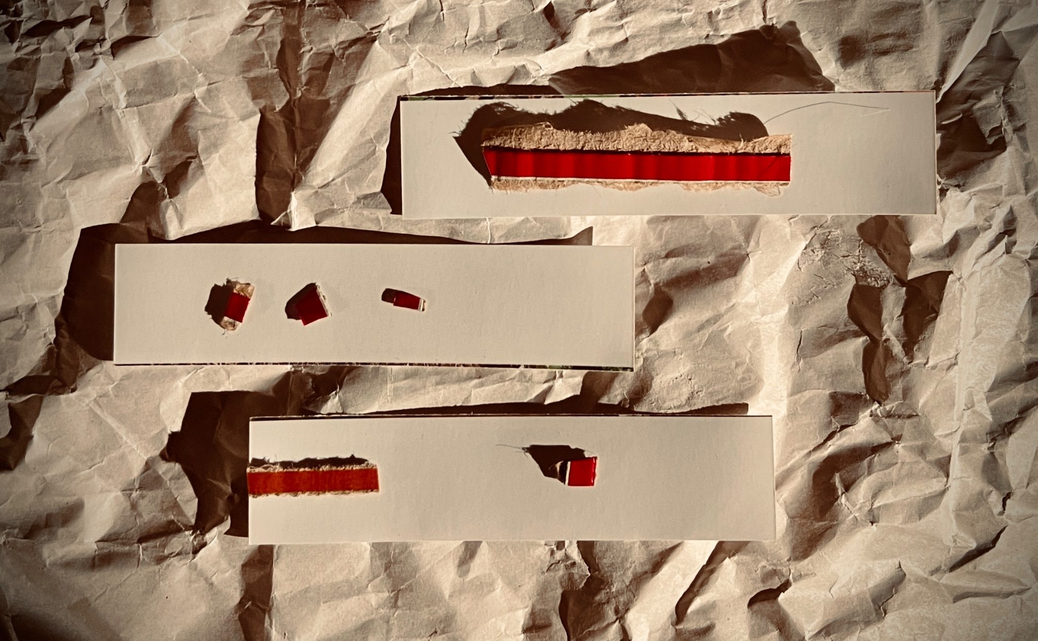 Three strips of white card are placed on a piece of crumpled newsprint paper. On top of the strips are pieces of the rip-strips from cardboard envelopes. Abstract composition.