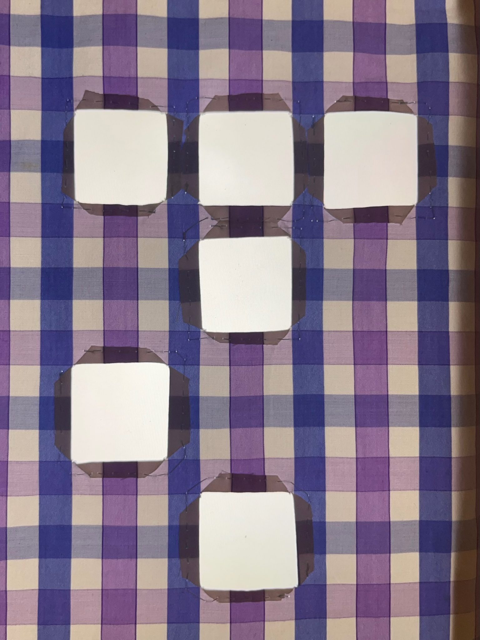 A pale purple and blue checked fabric backlit on a bright light box. 6 squares are cut out of the fabric, arranged irregularly