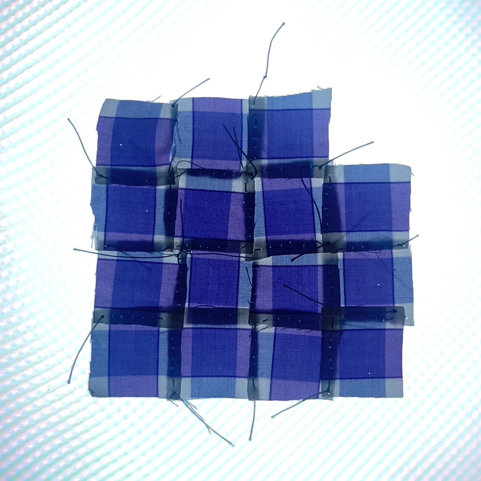 A patched piece of 15 tiny squares of fabric. Each is a section from a purple checked shirt, so the pieces form an irregular wobbly grid. The patch would be a square but the top right scrap is missing. Strands of thread stick out of the patch.