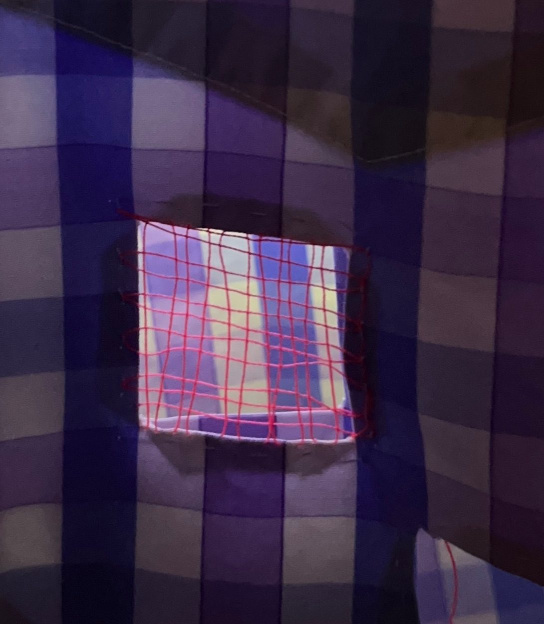 Close up photo of a square hole cut into the pocket of a cotton purple and white checked office shirt. Thin pink thread is woven across the hole making a very loose mesh. The fabric is backlit making the hole glow bright.
