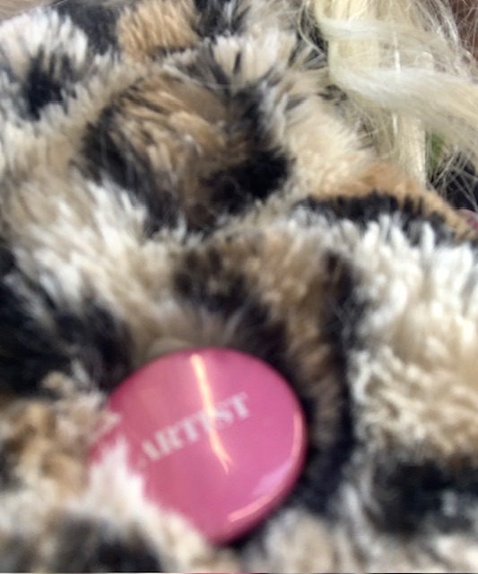 Colour photo of a pink badge with the word ARTIST in white letters and a yellow lightning flash across it. The badge is pinned to fake leopard fur. A curl of white hair is visible in the top right corner.