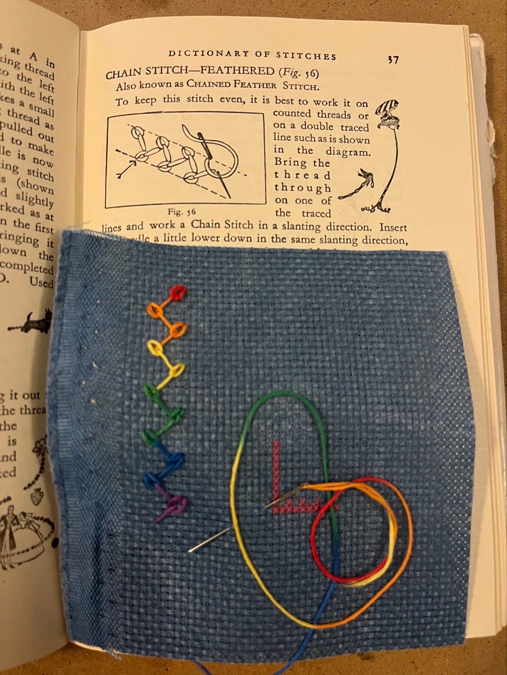 An open book with a piece of stitched blue fabric covering the bottom half of page 37. The top half of the page has a stitch diagram and text describing how to work the feathered chain stitch. On the fabric are small stitches in graduated rainbow colours, zigzagging up the left side. A loose strand coils next to the sewing.
