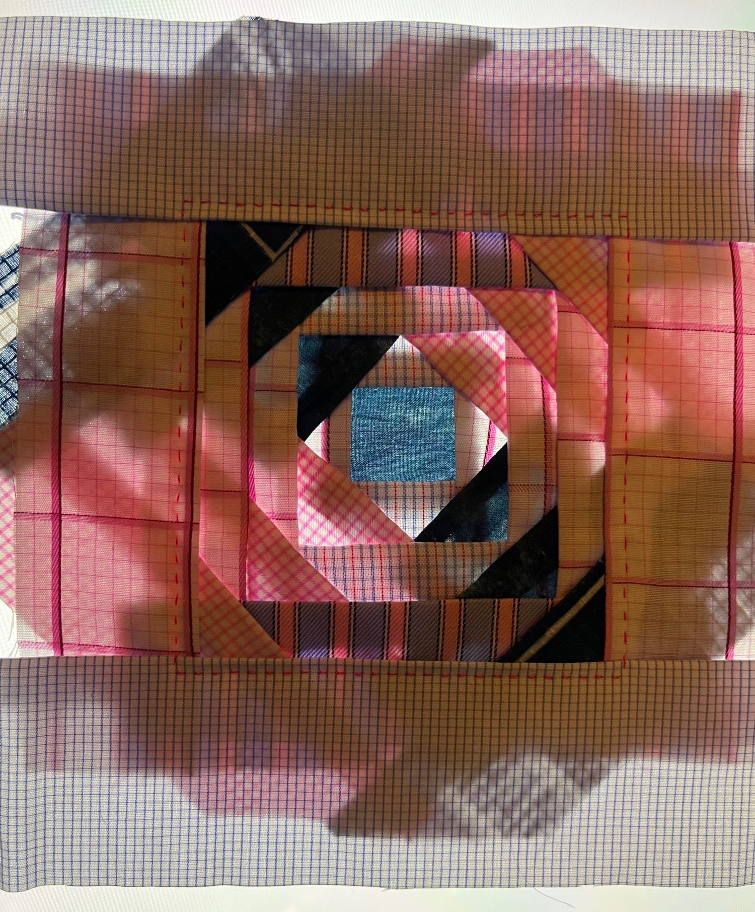 A roughly square textile piece backlit using a flat white light. The textile is mostly constructed from strips of formal office shirts in small blue or pink grids, pink and blue stripes, with some solid blue scraps. The backlighting shows dark strips where the scraps are layered and seamed together.