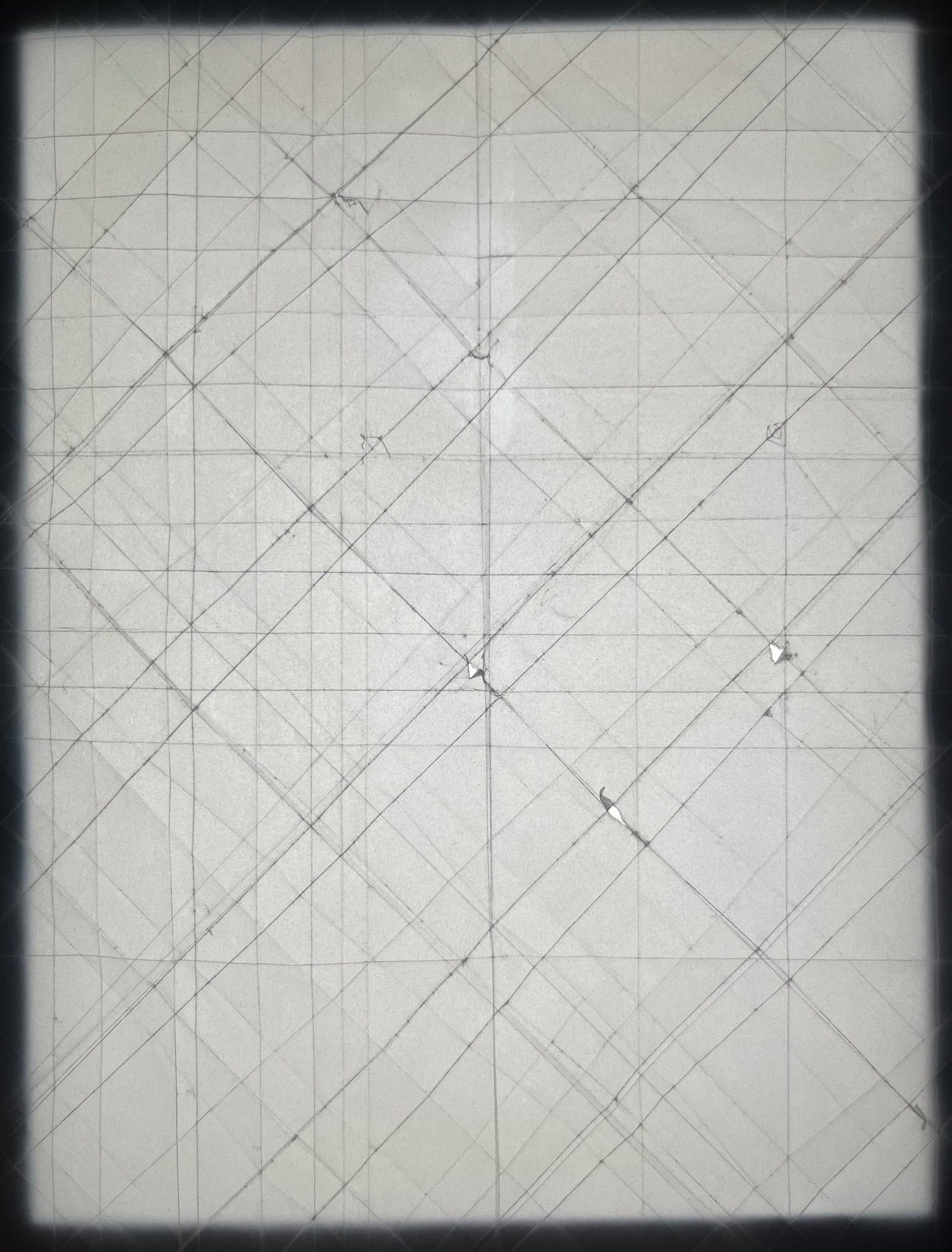 A sheet of tracing paper backlit with an even white light. Thin dark lines criss-cross the paper where it’s been folded and flattened. At some junctions there are tiny bright spots where the paper has torn.