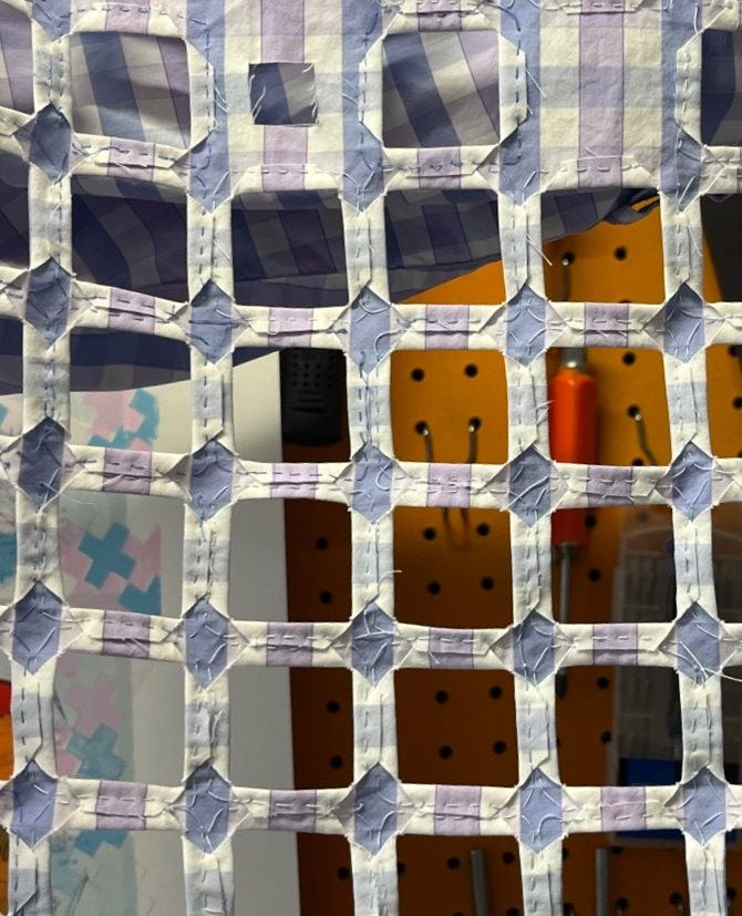 Close up on a rectangle of fabric that has a regular grid of square holes cut in it, neatly hemmed. It creates a pale purple trellis effect, allowing the orange wall behind to show through