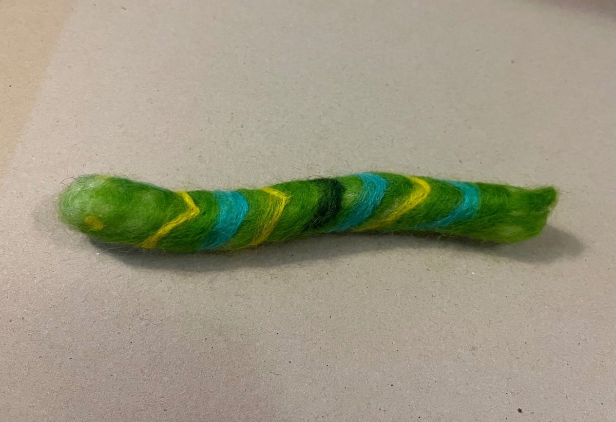A caterpillar made from needle felt on a buff card background. The caterpillar is chunky, bright green with blue and yellow chevrons running along its length.
