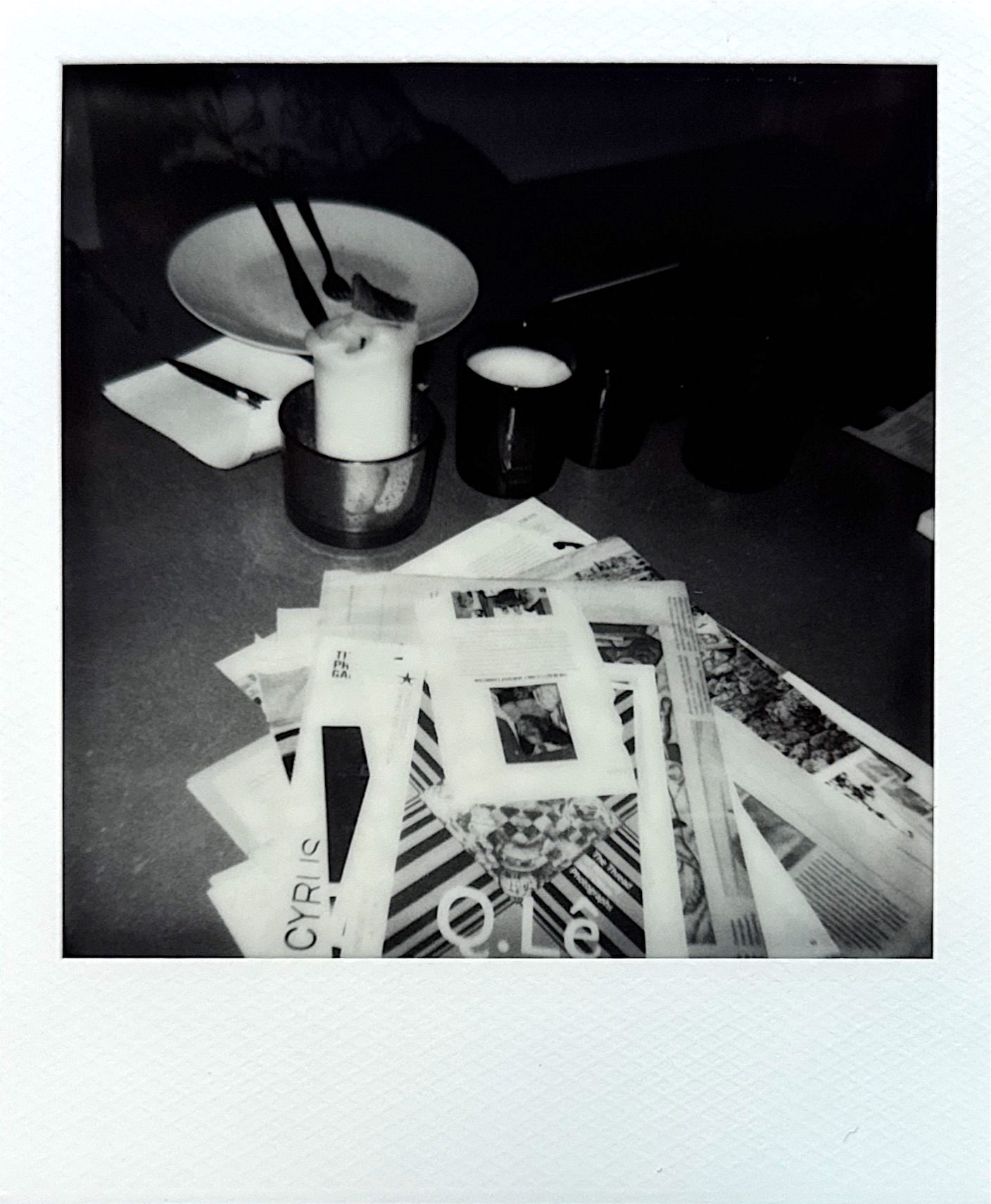 Black and white Polaroid image of papers dirty plate and candles on a table.
