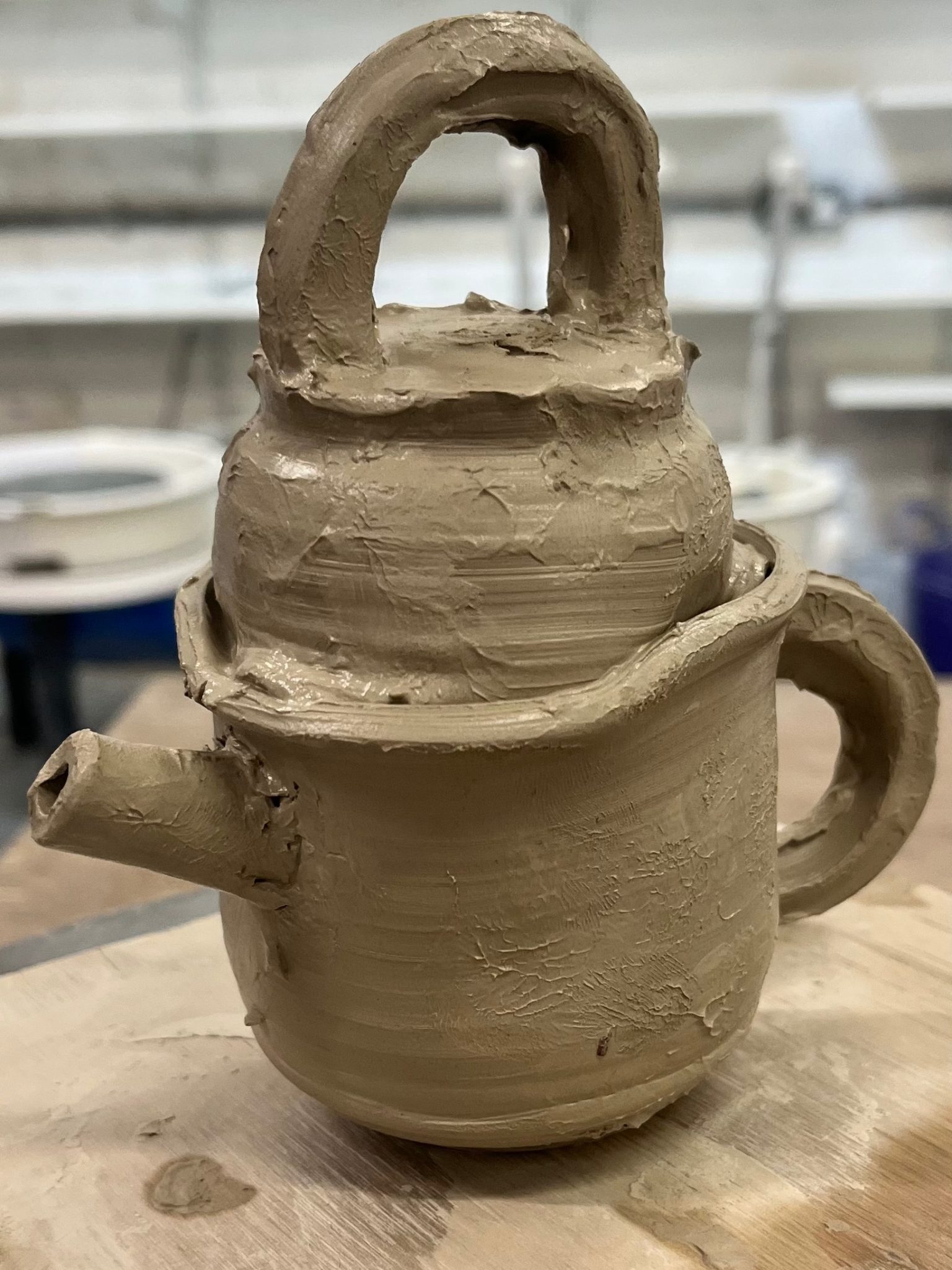 A clay teapot sits on a wooden board. It looks like 6 different pieces have been hastily thrown and squished. together.
