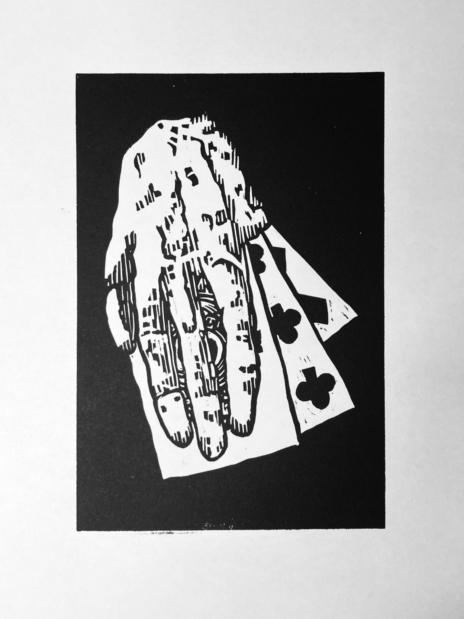 Black and white lino block print of a severed hand on top of three playing cards, the uppermost card being the ace of spades