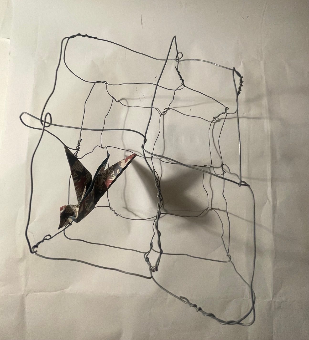 An origami bird in a wire cage (of sorts) with it’s shadow