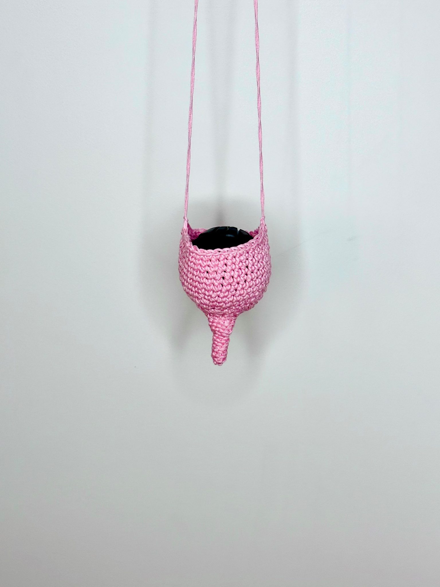 Photo showing a pink crocheted soft sculpture in a breast like shape, suspended from above against a white background with a black massage ball inserted inside