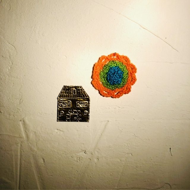 On a white wall I pinned a small house made of pushed aluminum, and a crochet work in the shape of a blue green orange sun