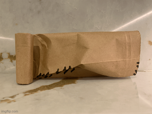 brown paper sculpture small with a curled column on one side and the rest of the sprawling paper on the other. small black marks have been drawn where the edges of the paper and the surface visible beneath meet.