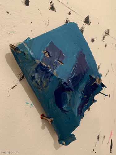 a small blue cardboard sculpture shaped like a quadrilateral whose diagonal fold is sealed with blue paint resting on two screws on a wall. wall is white with black paint marks on it.