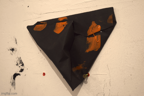 a triangular shaped black paper sculpture on a wall with the bottom on two screws coming from the wall. three orange brush marks of paint are visible across a line where parts of the paper meet due to being folded over.