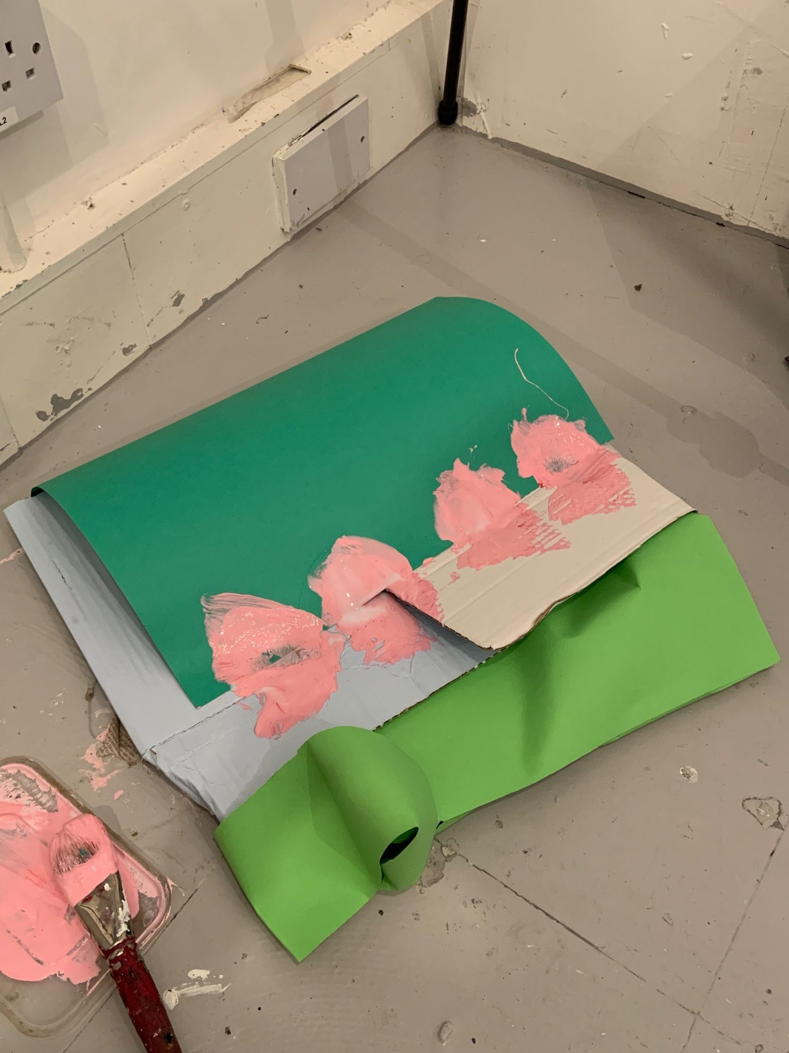A green piece of card stuck to white and blue cardbord pieces with splodges of pink paint, and a lighter green piece of paper on the opposing side. On the floor.