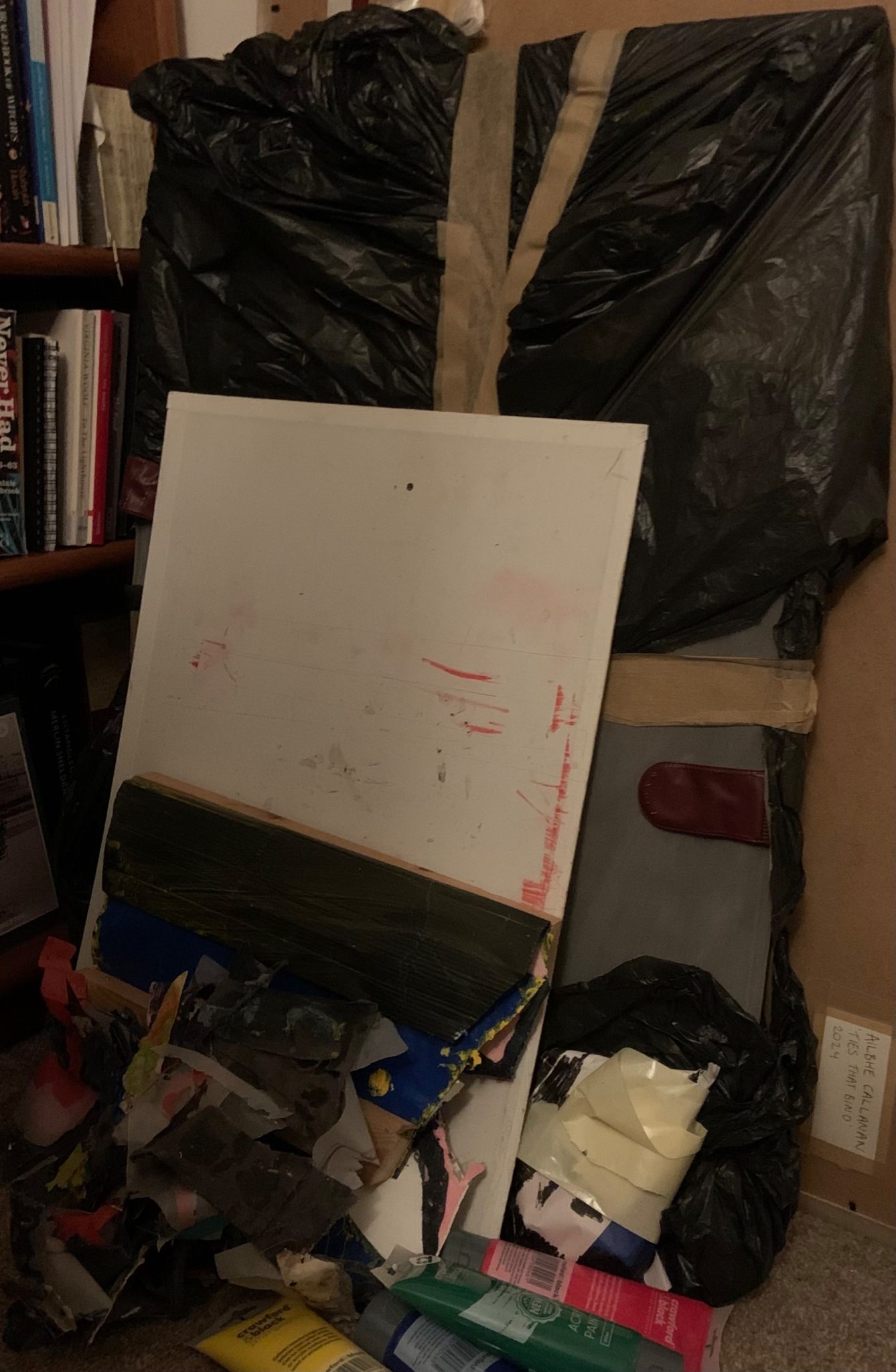 a stack of a frame, something in a bin, material on the floor, and four tubes of paint in the corner of a half lit room next to a bookcase