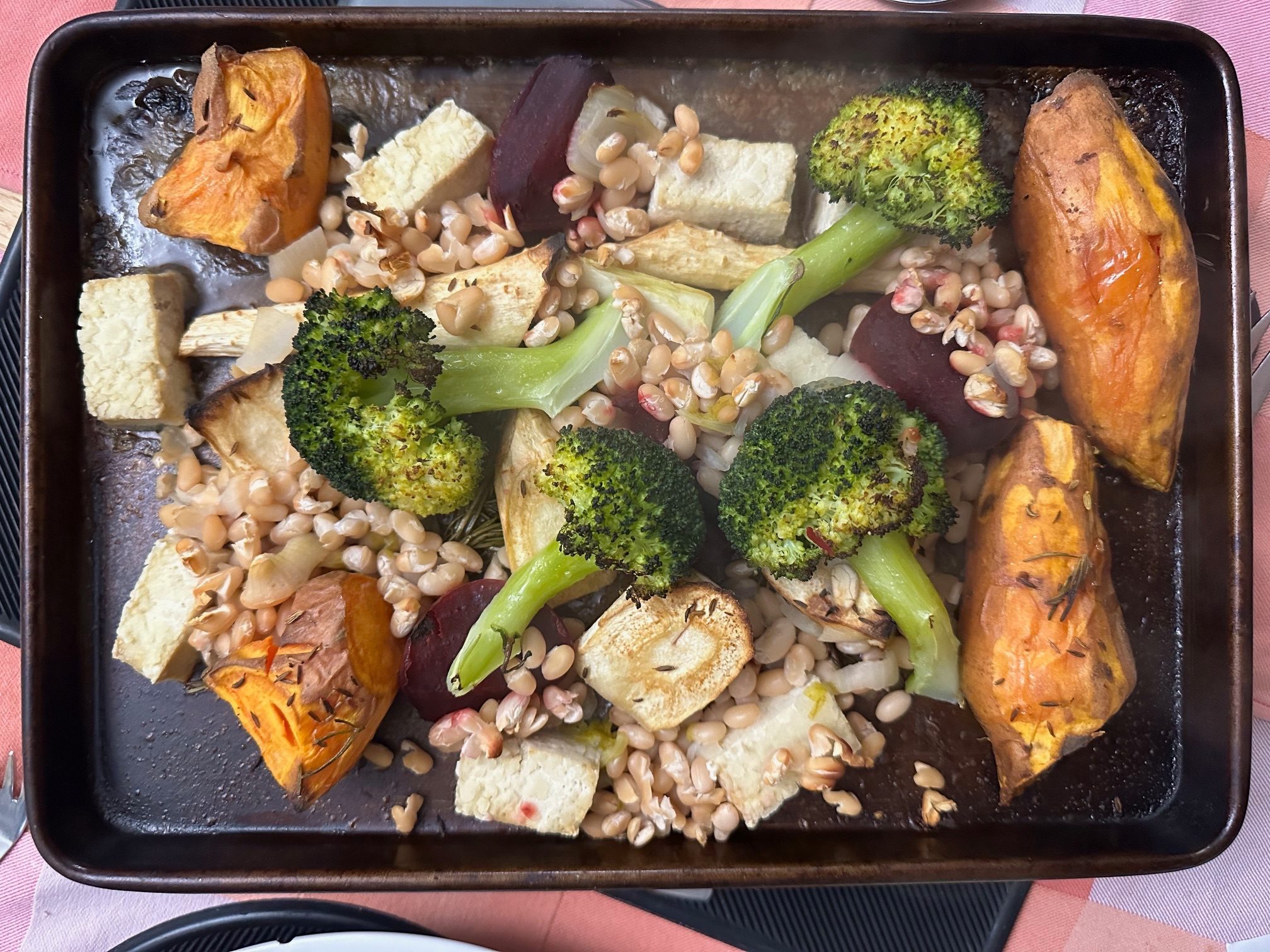 Photograph. Roasted vegetables of varied shapes and colours arranged  on black roastng tin