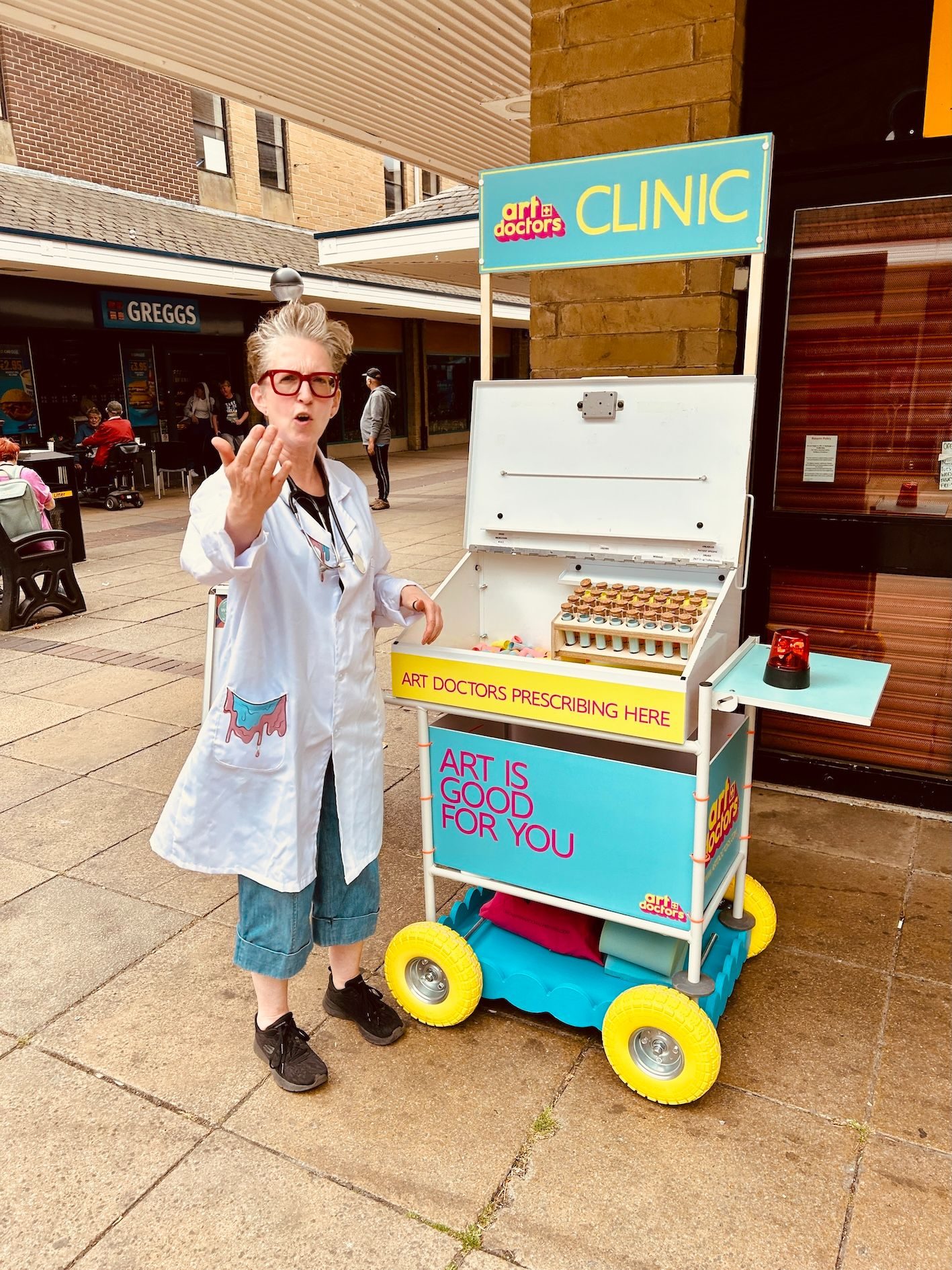 White woman in an Art Doctor branded coat standing next to a branded NHS dispensing trolley and encouraging passers by to come and talk to her
