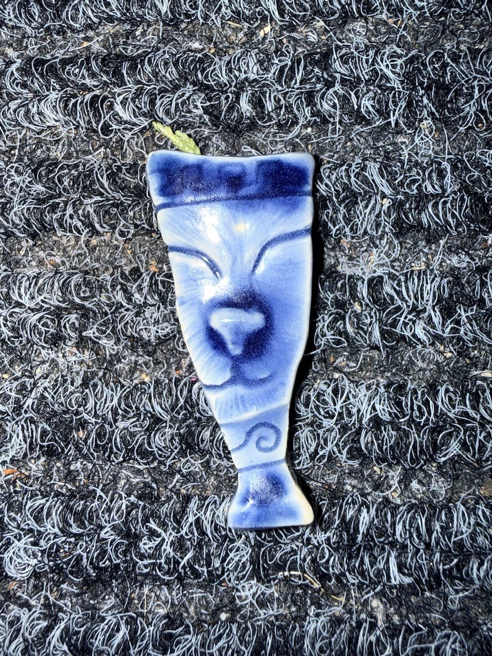 A vaguely vase shaped shard of ceramic with a close up of a cats face sculpted in to it. Top and bottom there is the typical greek vase swirls. It is glazed in blue and photographed on a door mat close up