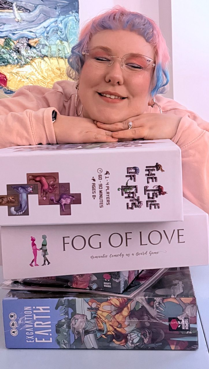 Picture of the artist with heavy make-up and styled hair, resting their head on the hands on-top of a stack of board games