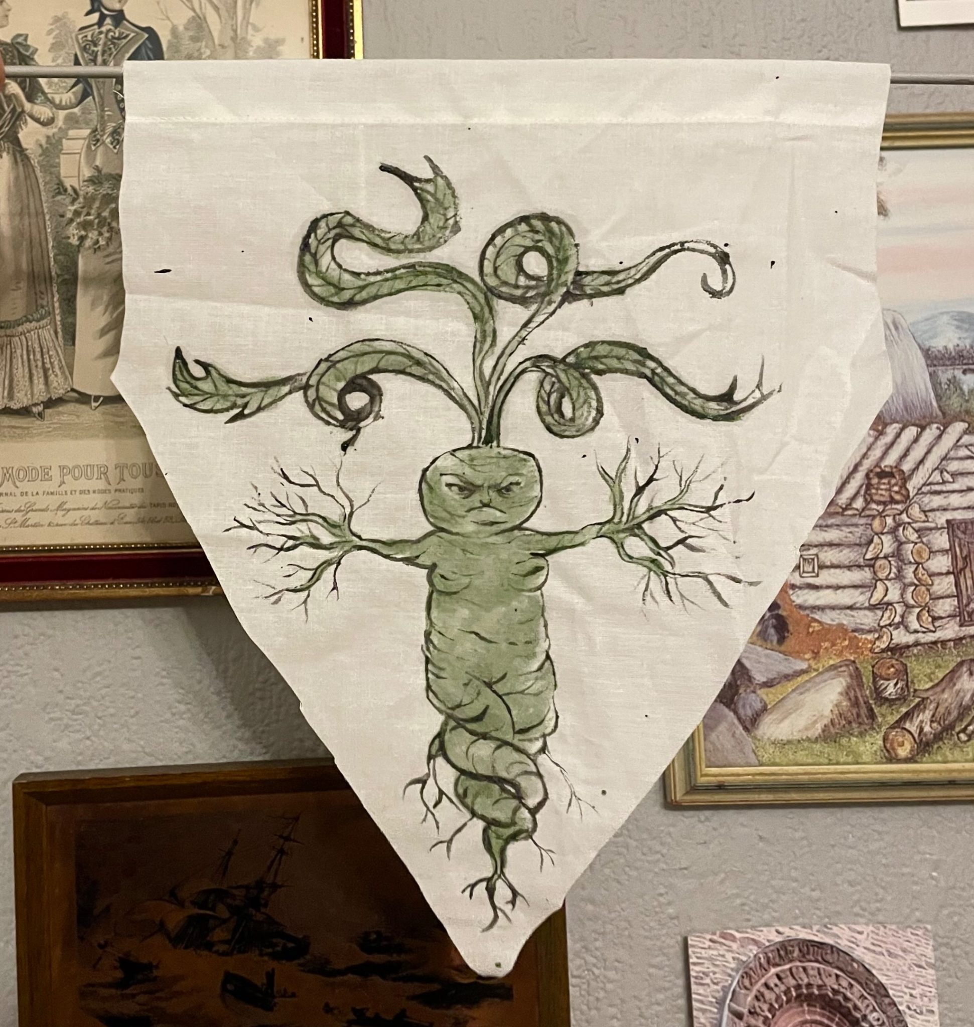 Mandrake woman painted onto a downpointed fabric triangle. She is spreading her roots and has an angry expression.