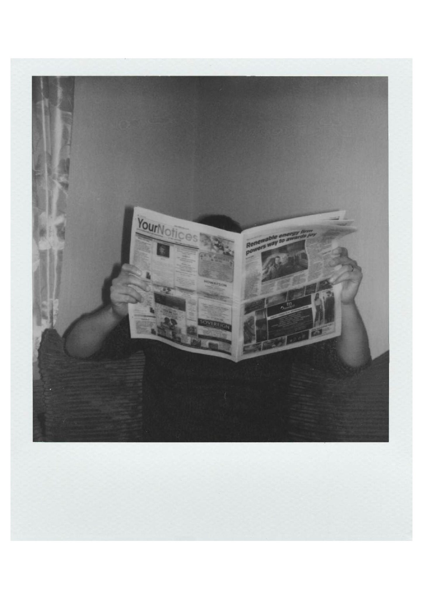 A black-and-white Polaroid photograph shows a seated person holding a wide-open newspaper that completely obscures their face. The headline 