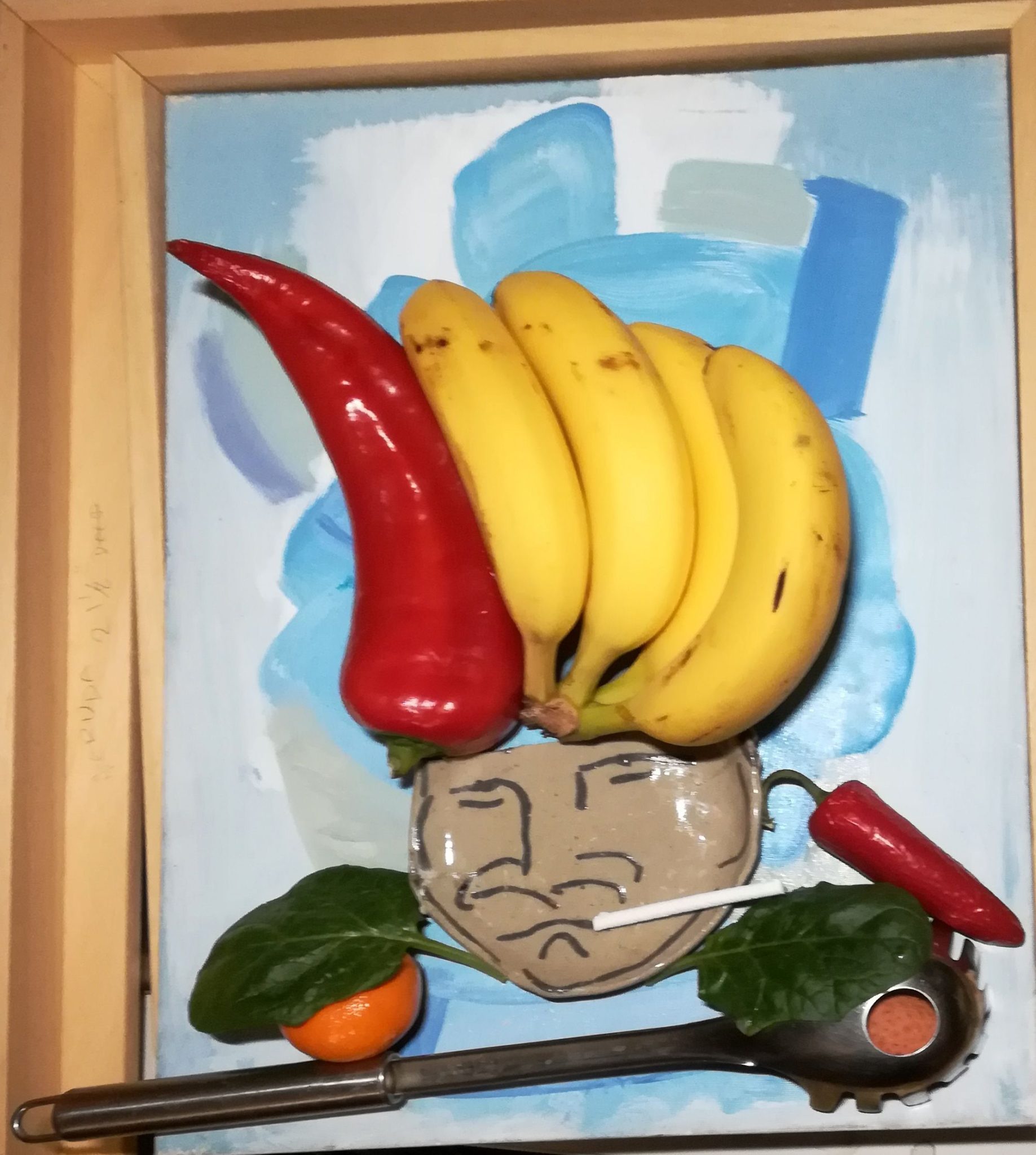 Broken small plate with grumpy face, bunch of bananas & Red pointy pepper for hair, chile pepper ear, pasta server  garnished with spinach leaves over tangerine and lychee for shoulders, candy cigarette in corner of mouth laid on canvas painting of blue abstract headless fat figure,with 2 frames