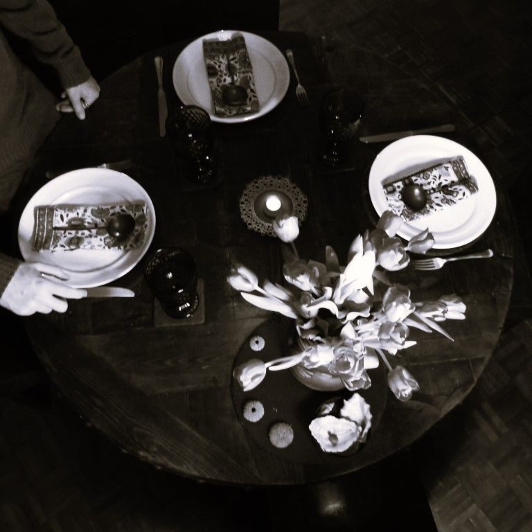 Overhead photograph of setting the dinning table - round table with flowers and a candle, three places being set by son. Black and white photo
