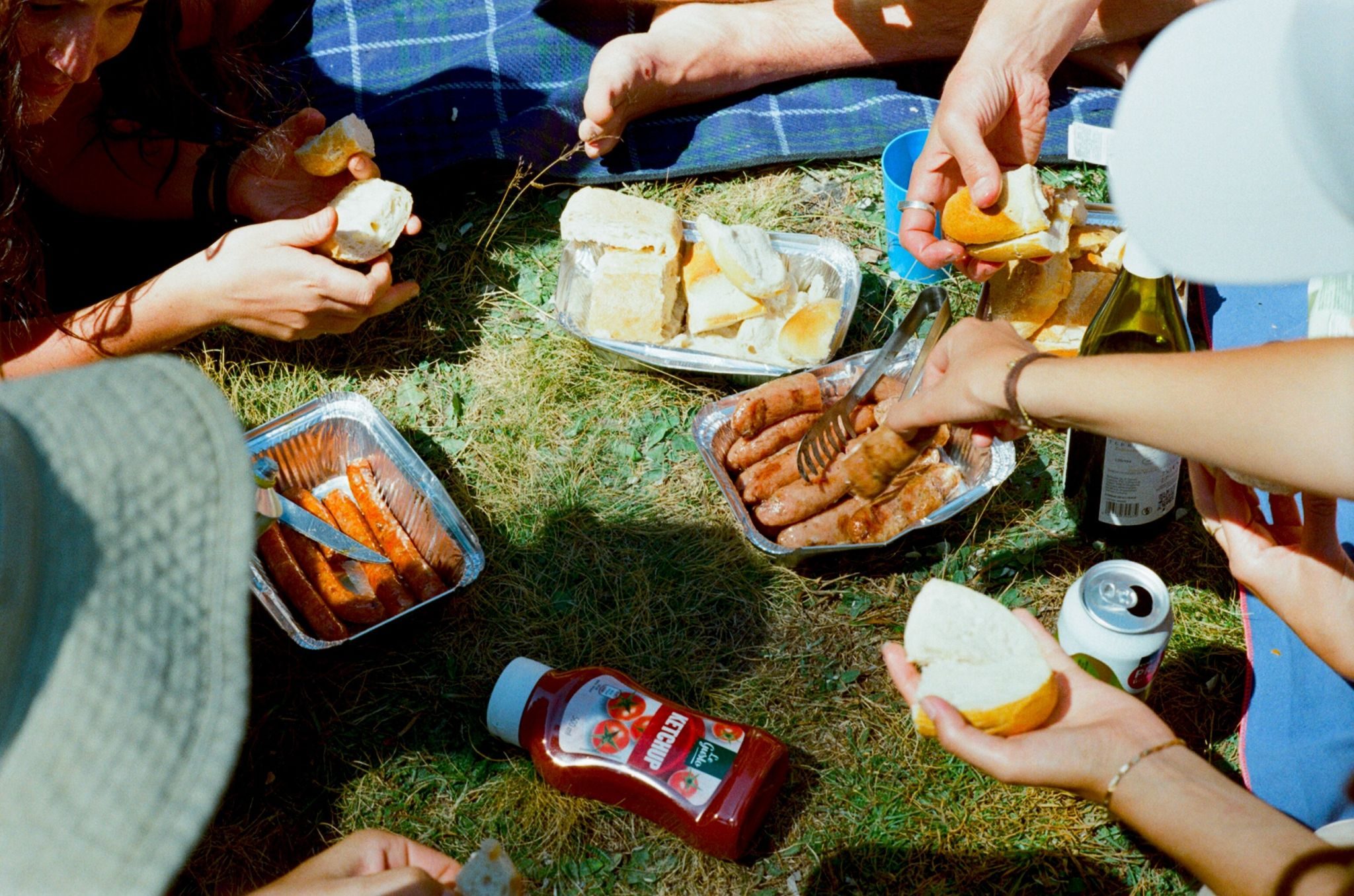 A close up shot of hands grabbing food at a picnic - details of sausages, bread and ketchup bottles lying on the ground.