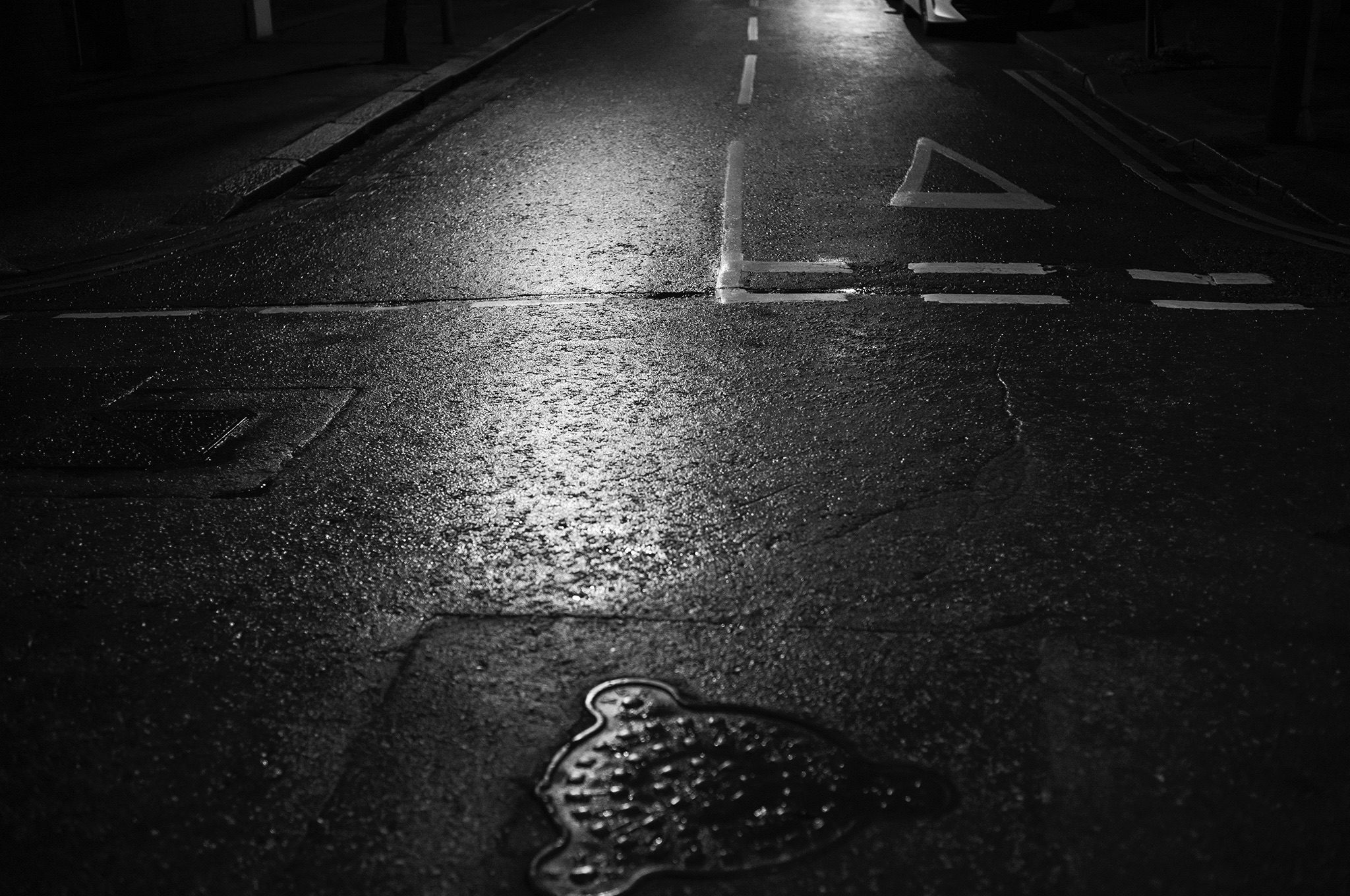 Black and White image of a deserted street at night with manhole covers and street markings on it. Dark and foreboding