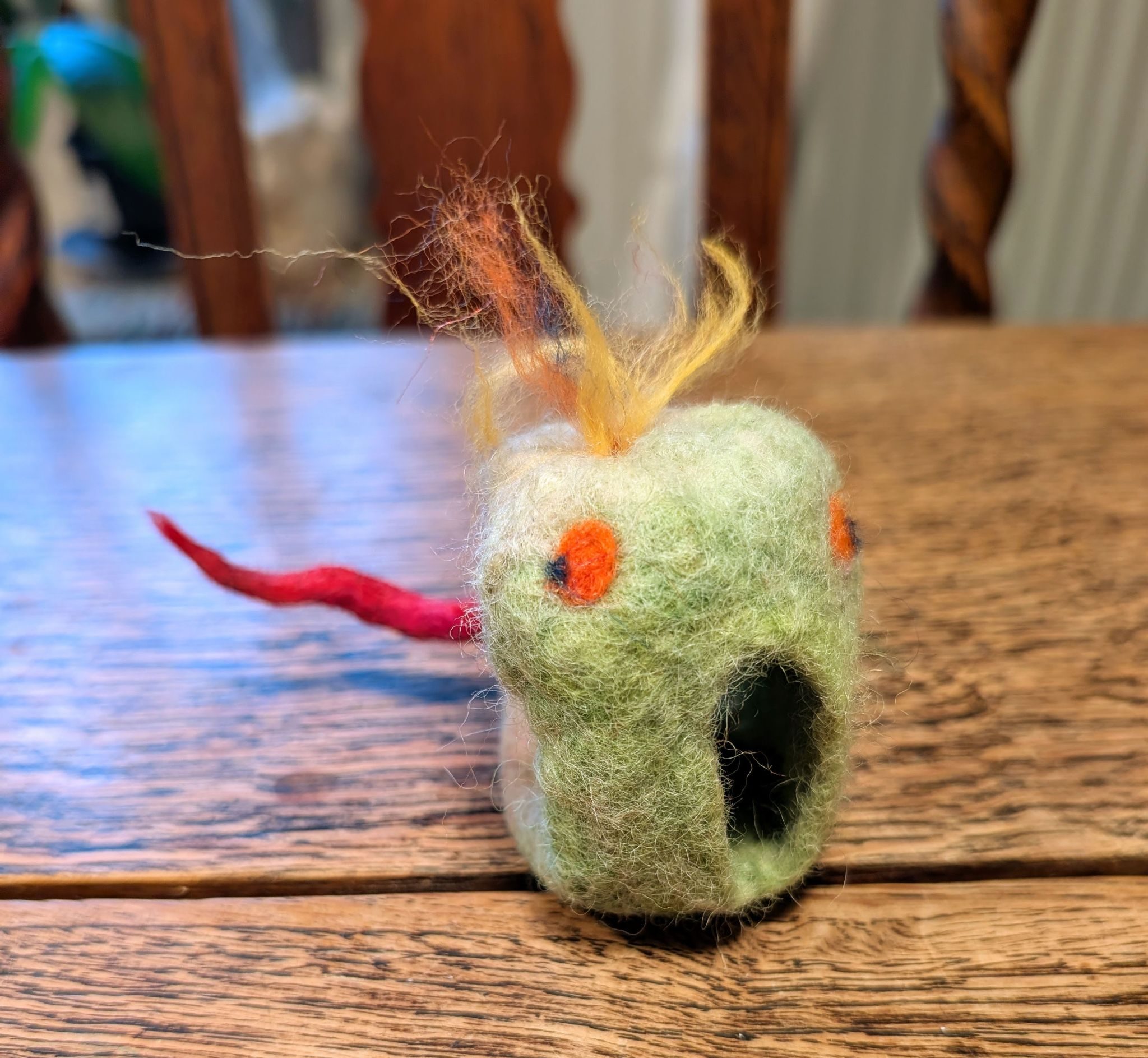 A small felted figure made from light green wool, with two round orange circles on the front that resemble eyes. It has a mouth like opening that has a stunned appearance and a tuft of yellow‑orange wool attached to the top. A red, twisted wool piece extends from the back like a tail.