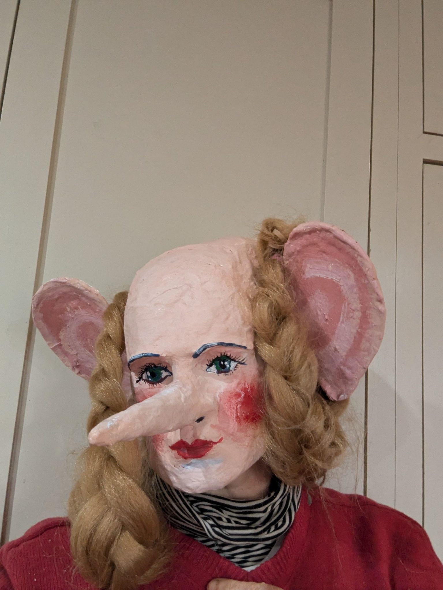 A woman wearing a papier mache mask with a long nose, big ears and blond braids.