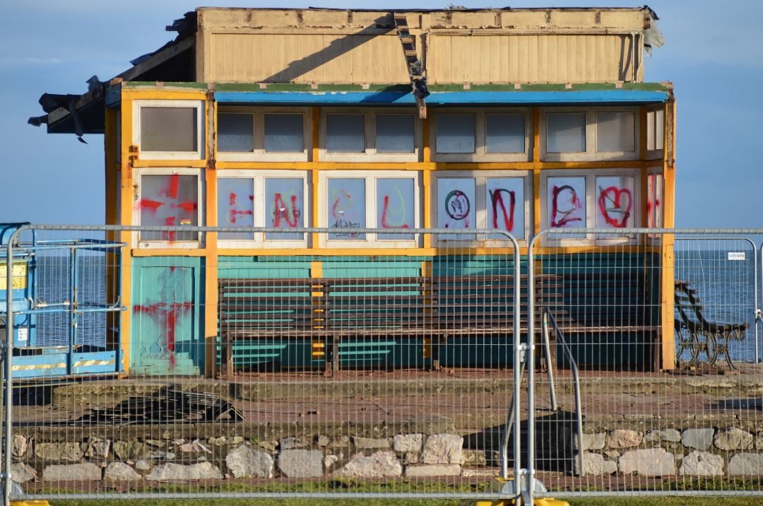Battered partially deconstructed beach shelter, which is graphited with red scrawls and half rubbed away crosses. Temporary metal fenceing in front and a calm sea and clear sky behind.