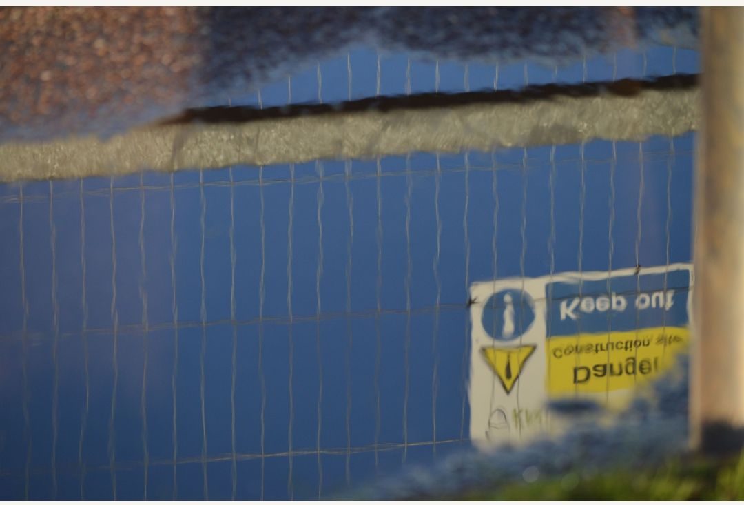 Sign saying danger keep out, as seen reflected in a puddle with blue sky behind