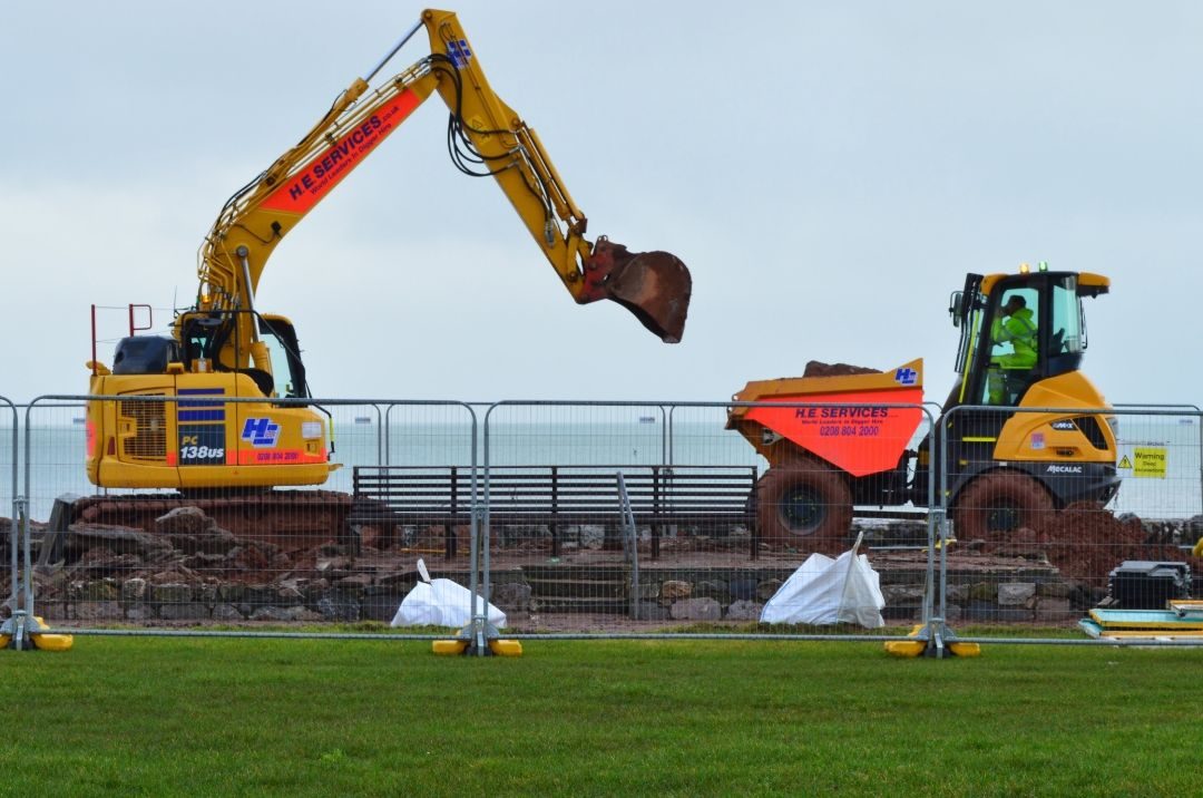 Colour photo of an excavator poised to pick up its next load of soil at a fenced construction site.
