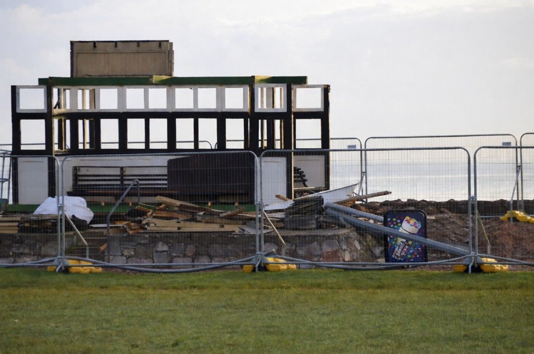 A partially demolished seaside shelter behind fencing. Free standing ice-cream advert stands amongst the deconstructed pieces.