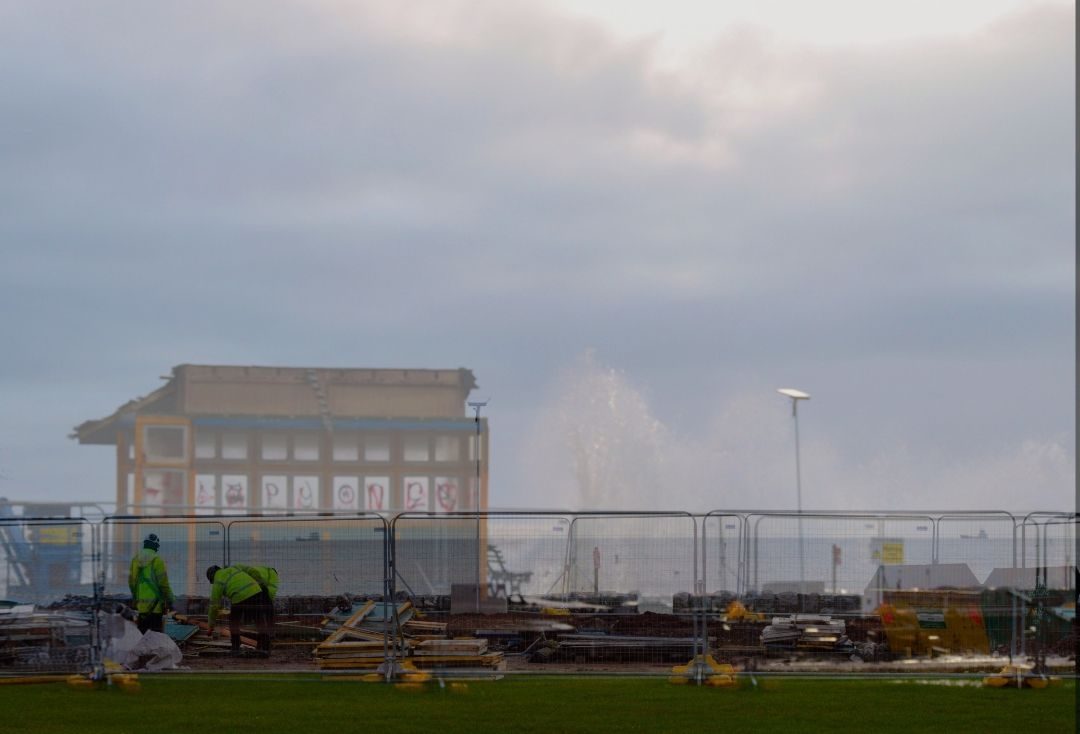 A faint, ghostlike image of a former seaside shelter overlaps workers clearing debris behind fencing as waves crash under a grey sky.