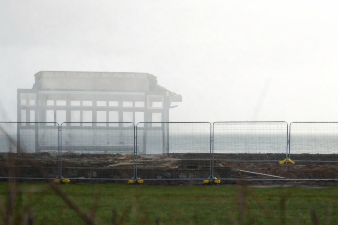 Battered partially deconstructed beach shelter, which is graphited with red scrawls and half rubbed away crosses. Temporary metal fenceing in front and a calm sea and clear sky behind.
