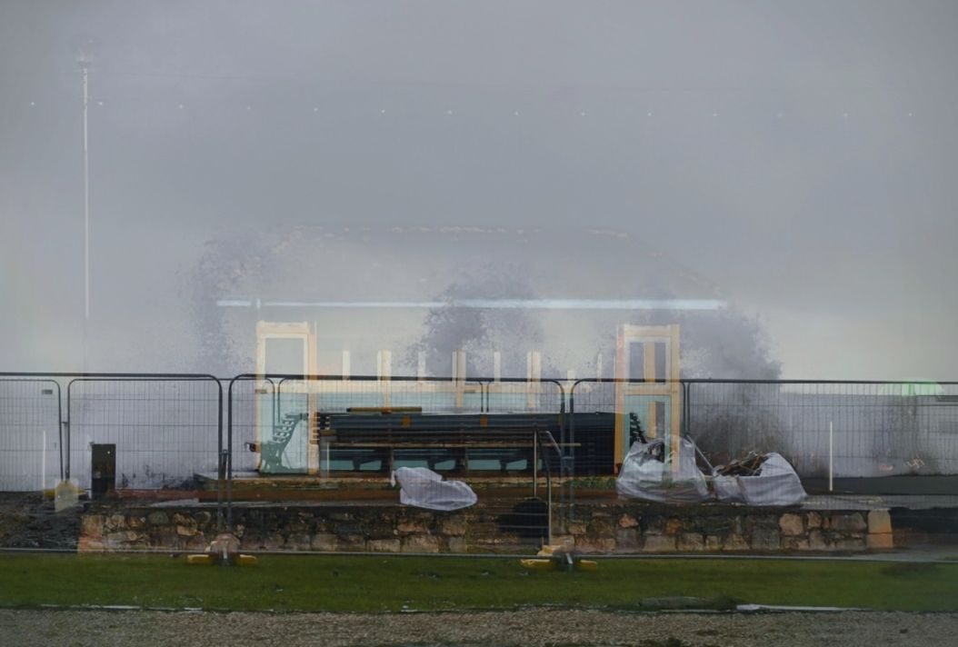 Remains of a largely demolished beach shelter. Large waves crashing over the sea wall behind. The ghostly image of the original shelter visible on top