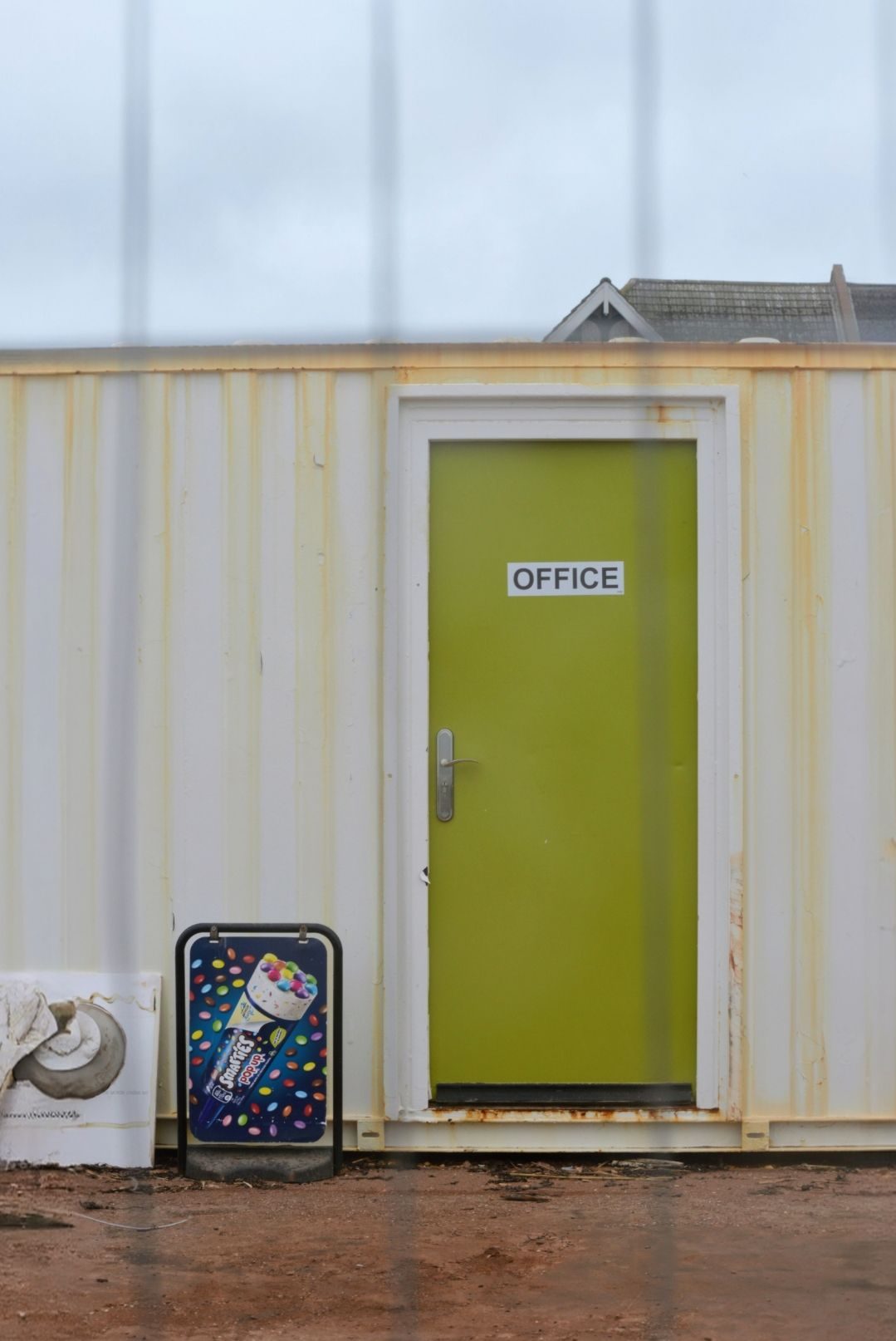 Partial view of a shipping container with a green door marked office next to which is an ice-cream  advert