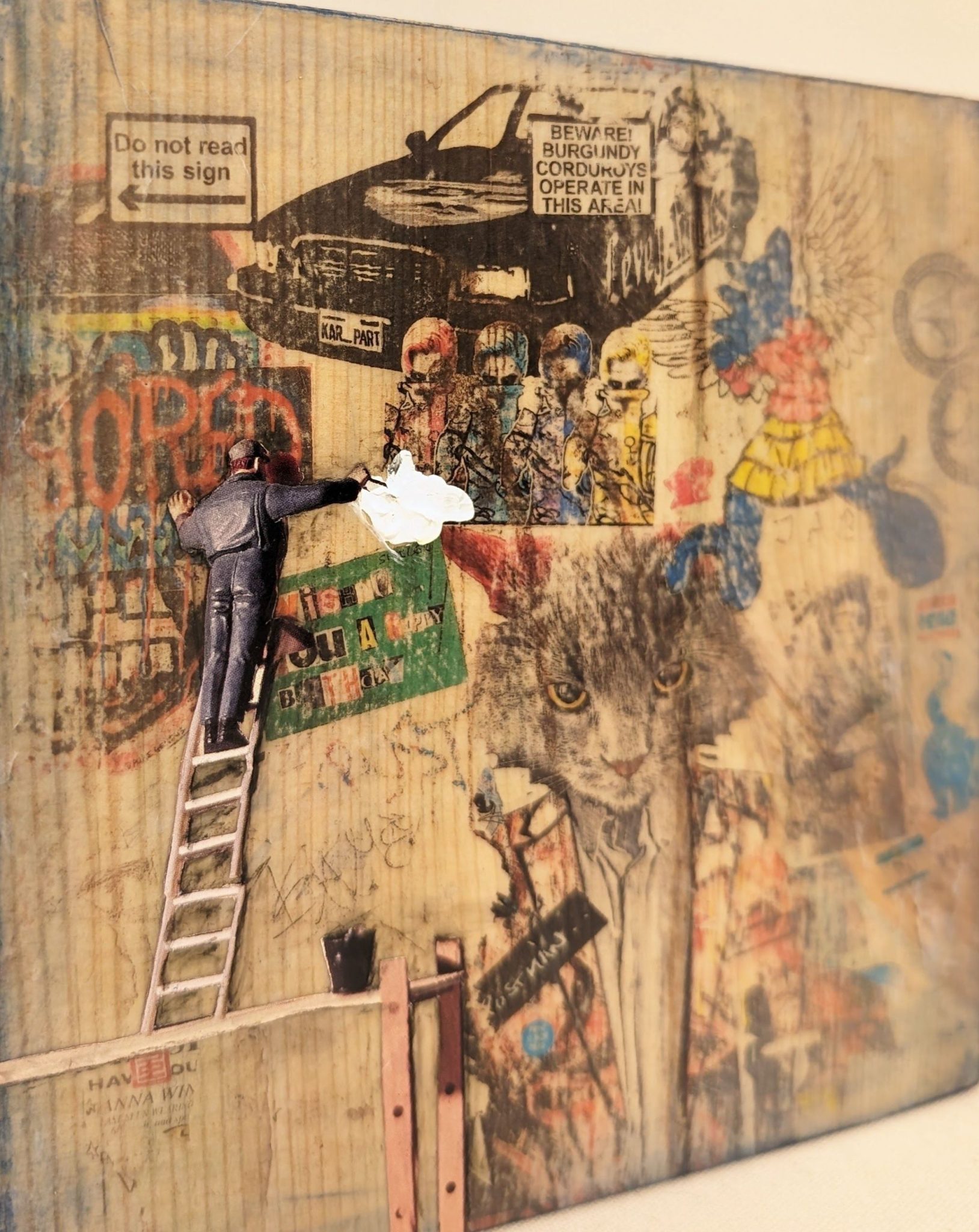 The artwork is a dense, playful collage combining flat images, graffiti, and a tiny three‑dimensional figure of a man on a ladder. Around him are printed characters including hazmat‑suit figures, a standing cat in a coat with a “POSTMAN” badge, and a cartoon winged figure. Scattered signs and posters add to the chaotic, street‑wall atmosphere.