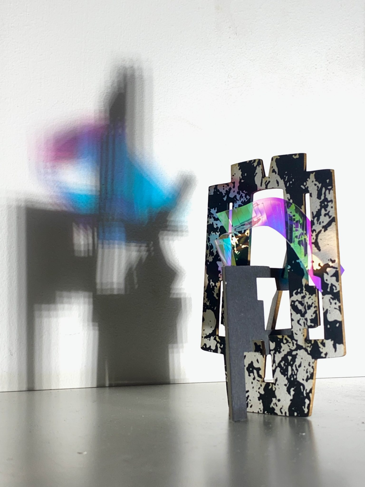 This small maquette is made from laser-cut, screen-printed card, cut and scored before being layered upright into a shallow architectural form. Rectangular openings and a strip of dichroic film allows light to pass through, casting repeating, colourful, shifting shadows behind the work.