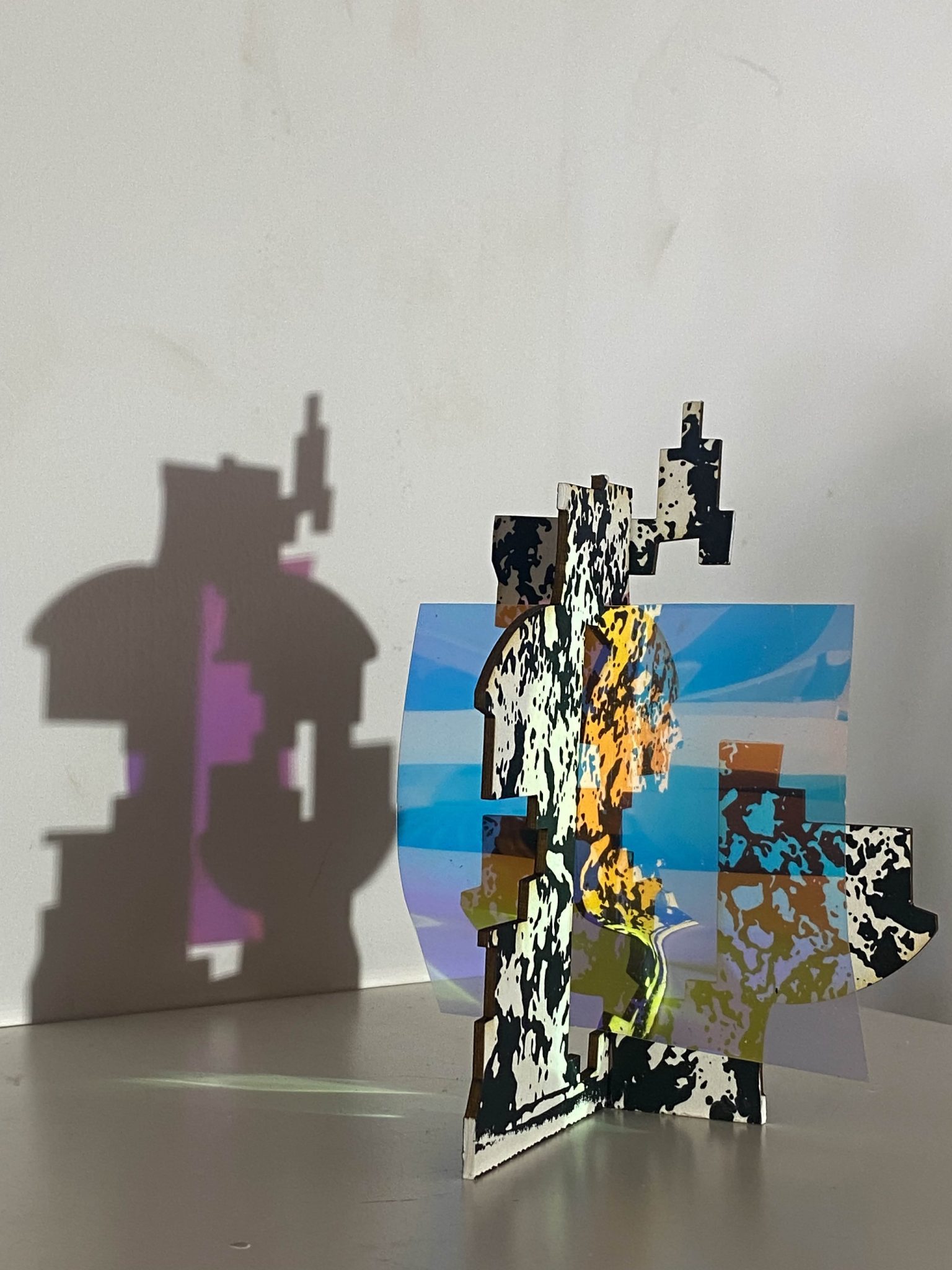 This small maquette is made from laser-cut, screen-printed card, cut and scored before being layered upright into a shallow architectural form. Rectangular openings and a strip of dichroic film allow light to pass through, casting repeating, colourful, shifting shadows behind the work.