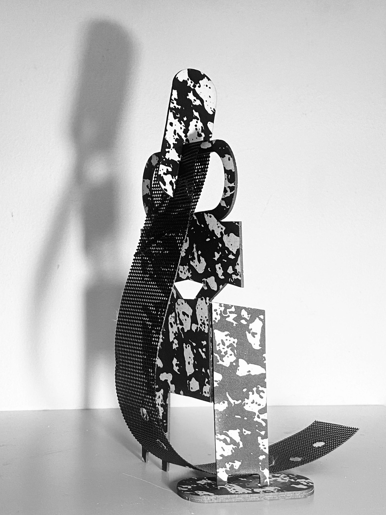 This work is a small maquette made from laser-cut card, presented upright on a flat surface. It is constructed from a flat screen printed card plane that is cut and scored, then layered vertically to form a shallow, architectural structure with rectangular openings. A strip of black mesh has been added. Light passes through the openings, creating overlapping shadows that repeat and shift the internal shapes onto the surface behind the work.