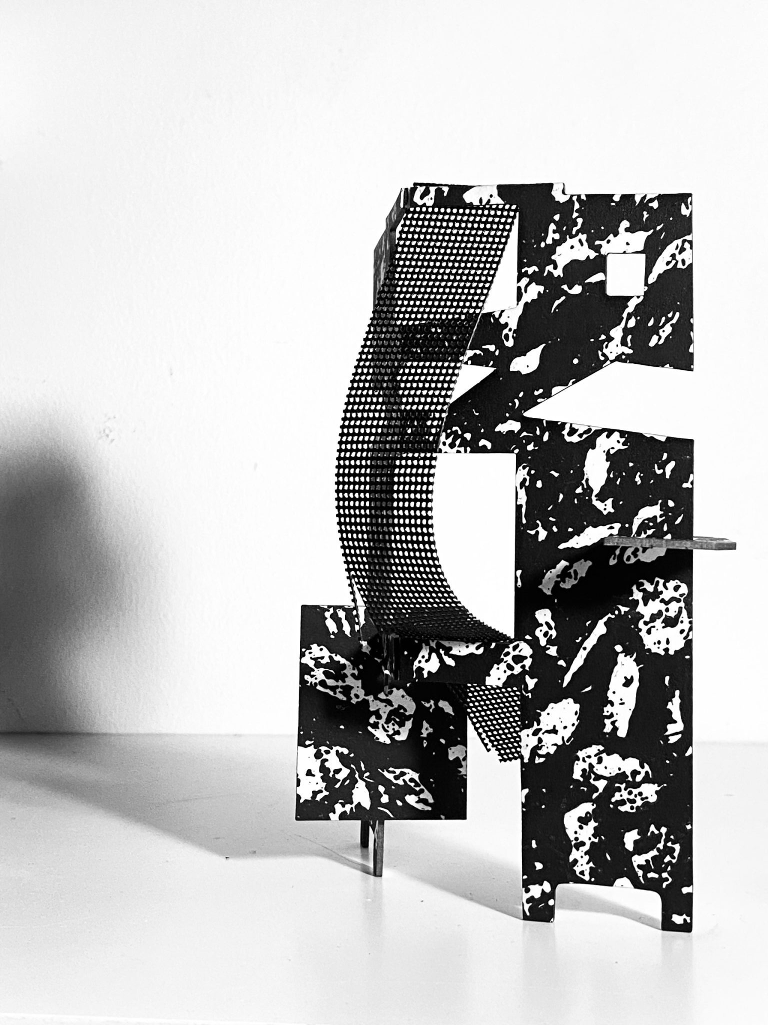 This work is a small maquette made from laser-cut card, presented upright on a flat surface. It is constructed from a flat screen printed card plane that is cut and scored, then layered vertically to form a shallow, architectural structure with rectangular openings. A strip of black mesh has been added. Light passes through the openings, creating overlapping shadows that repeat and shift the internal shapes onto the surface behind the work.