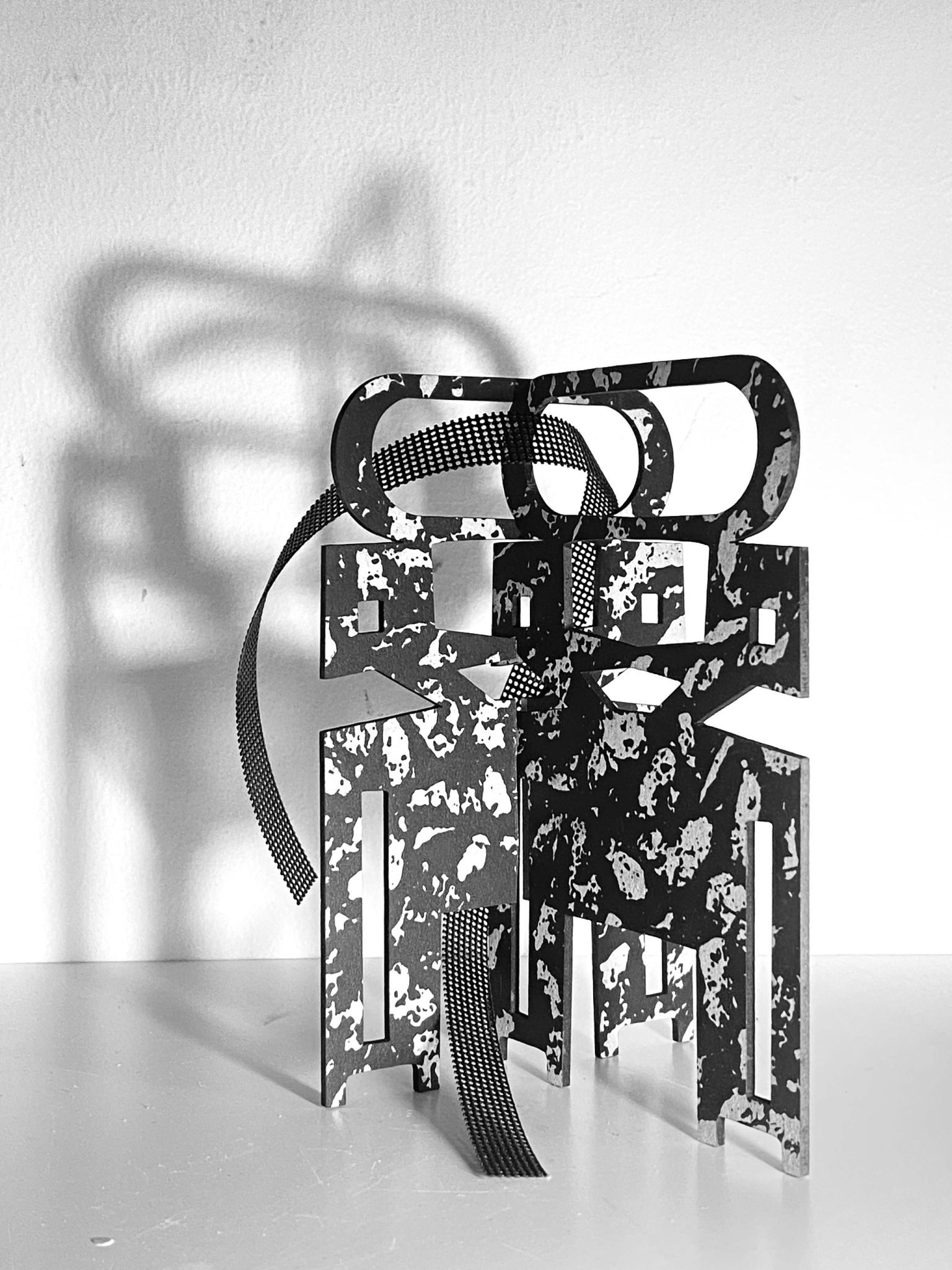 This small maquette is made from laser-cut, screen-printed card, cut and scored before being layered upright into a shallow architectural form. Rectangular openings and a strip of black mesh allow light to pass through, casting repeating, shifting shadows behind the work.