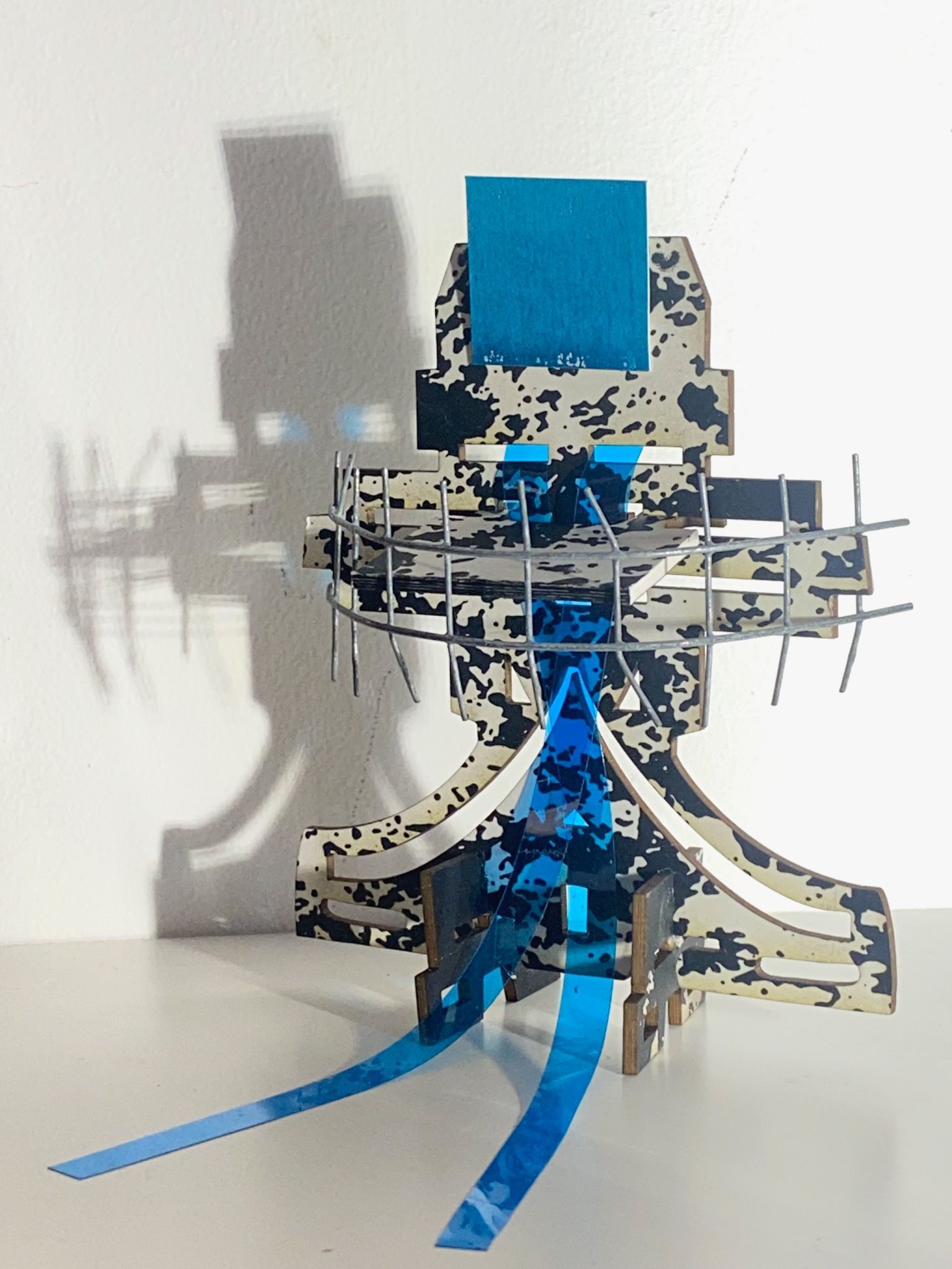 This small maquette is made from laser-cut, screen-printed card, cut and scored before being layered upright into a shallow architectural form, its front surrounded by curved wire mesh. Rectangular openings and a strip of blue film allow light to pass through, casting repeating, shifting shadows behind the work.