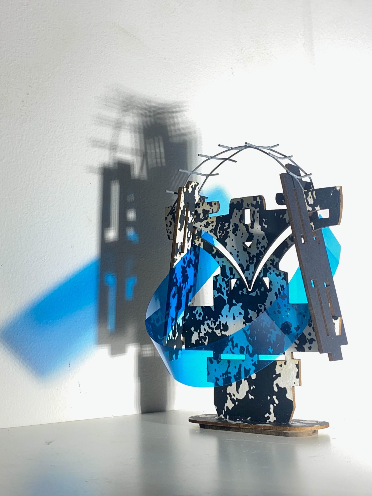 This small maquette is made from laser-cut, screen-printed card, cut and scored before being layered upright into a shallow architectural form, its front surrounded by curved wire mesh. Rectangular openings and a strip of blue film allow light to pass through, casting repeating, shifting shadows behind the work.